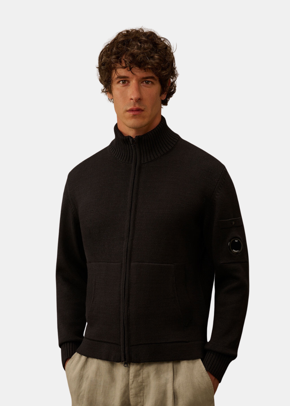 Mannequin homme de trois-quarts face portant le cardigan en maille de coton compact marine C.P. Company fermé, associé à un pantalon beige, mettant en valeur le col montant et le détail de la lentille noire