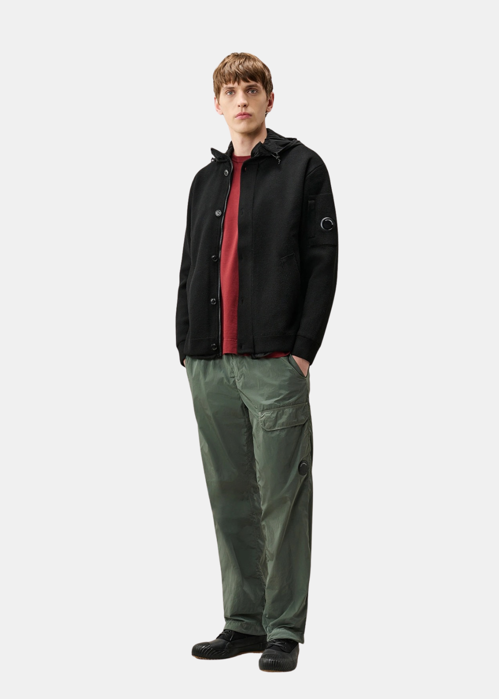 Silhouette complète d'un mannequin portant le cardigan C.P. Company noir ouvert sur un t-shirt bordeaux avec un pantalon parachute vert et des baskets noires