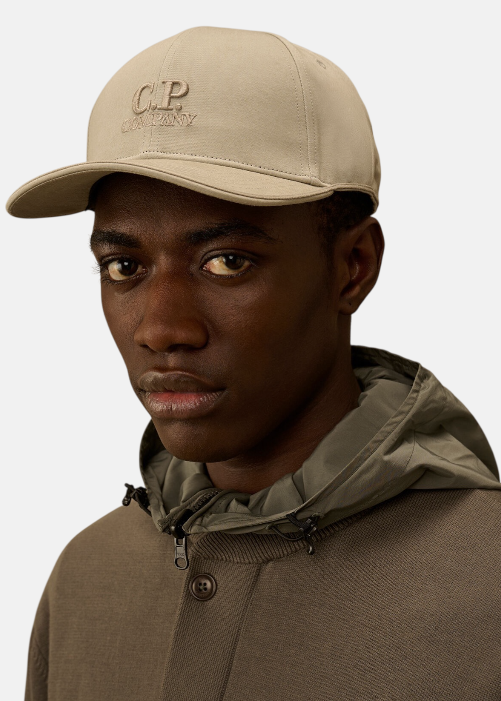 Gros plan sur le visage d'un mannequin noir de face portant la casquette C.P. Company beige sur fond uni blanc