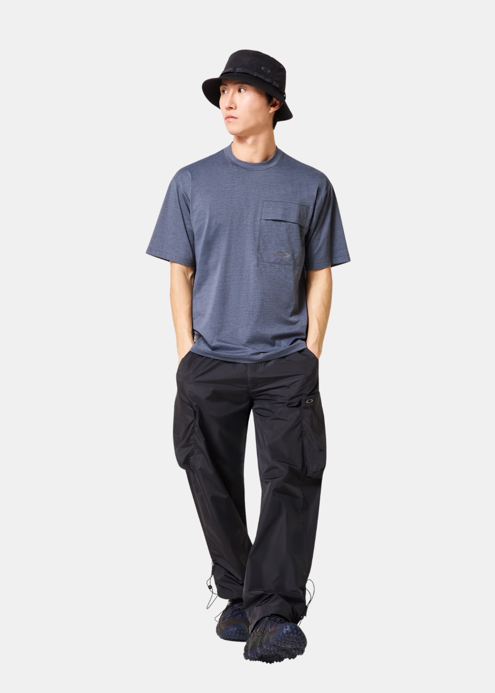 Homme portant un t-shirt bleu chiné Oakley, un pantalon cargo noir, un chapeau bob et des chaussures de sport.