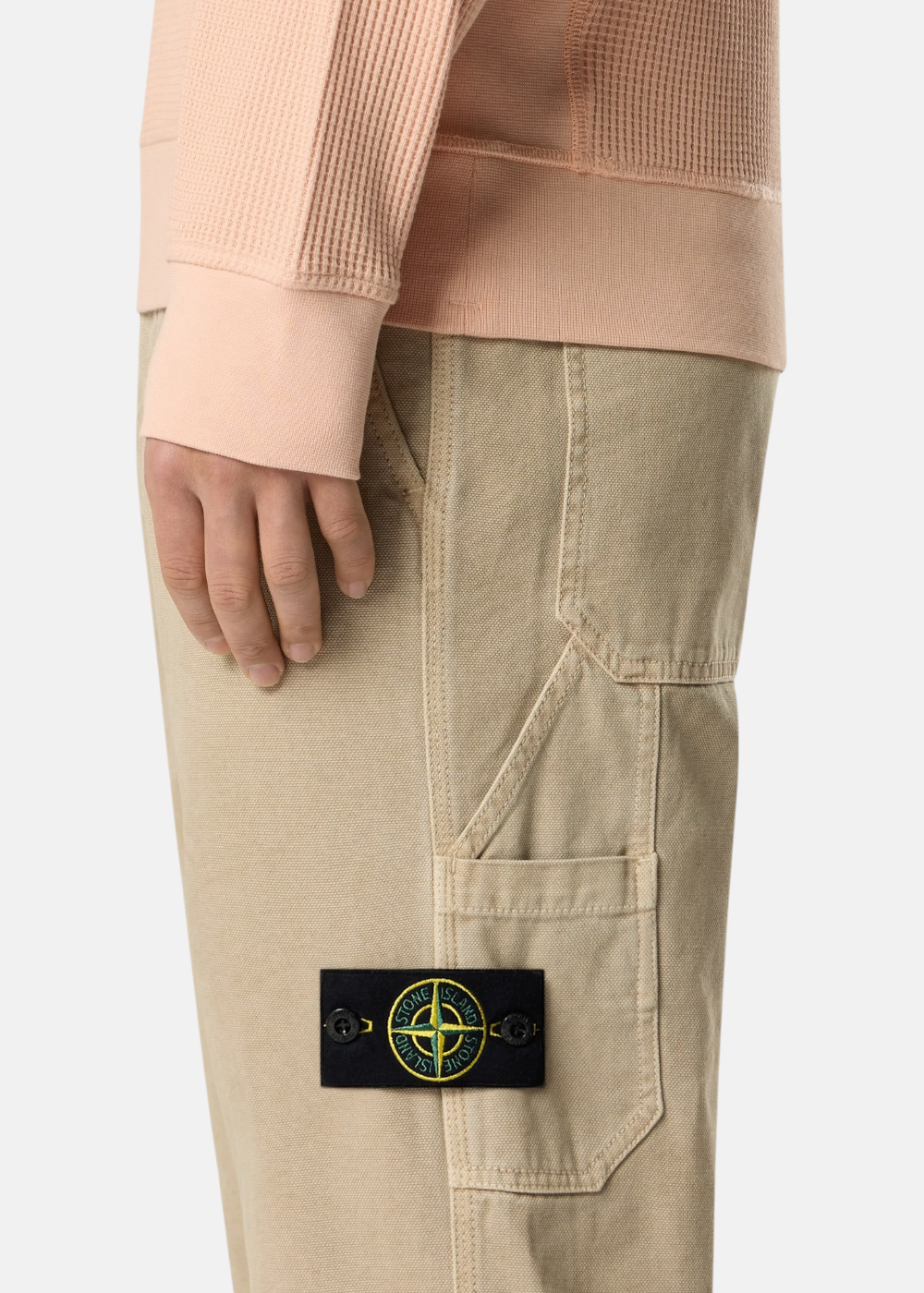 Gros plan sur le badge Stone Island amovible sur la poche latérale d'un pantalon beige en coton.