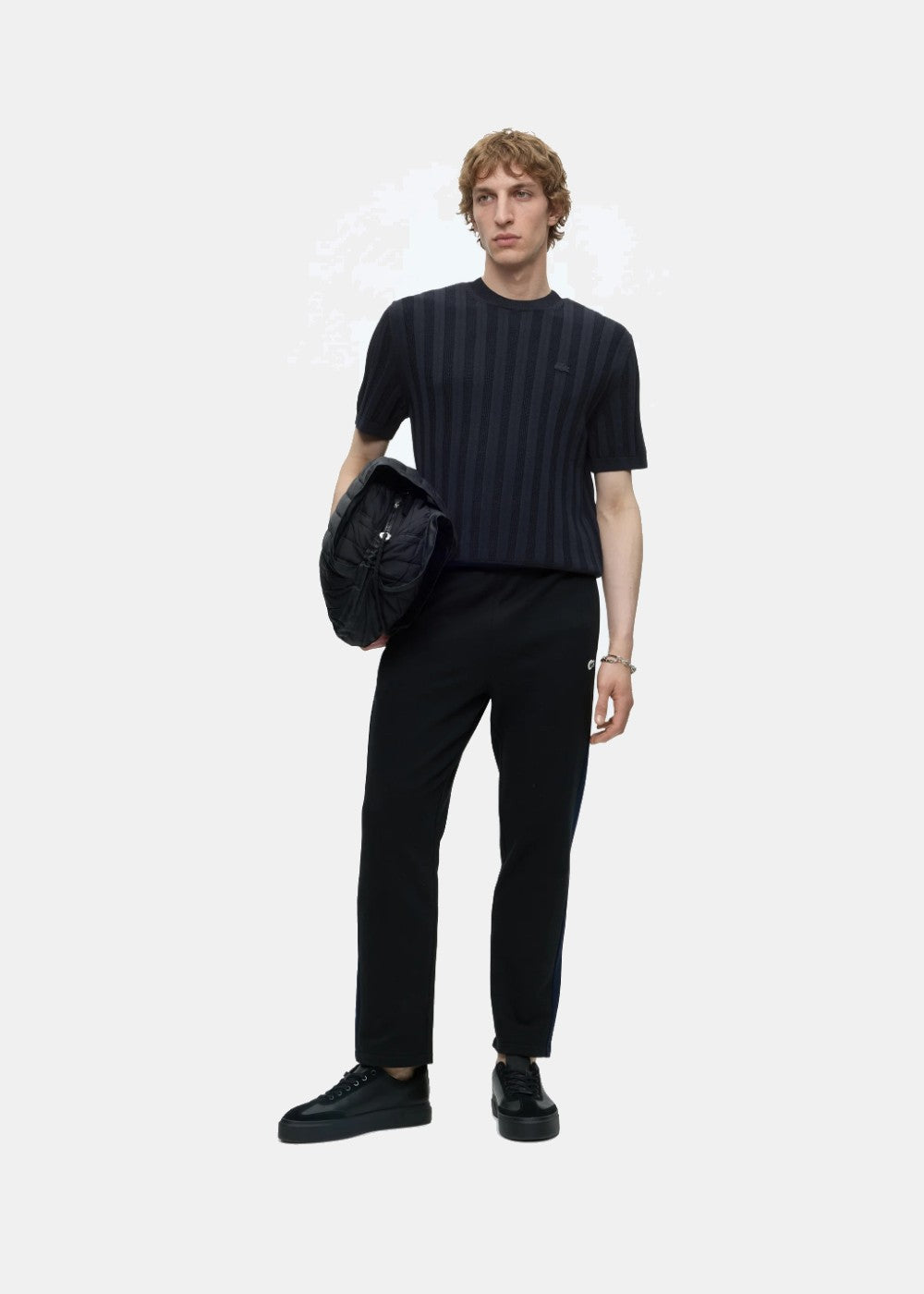 Porté en pied d'un polo Lacoste marine en maille côtelée associé à un pantalon noir, des sneakers noires et un sac matelassé.