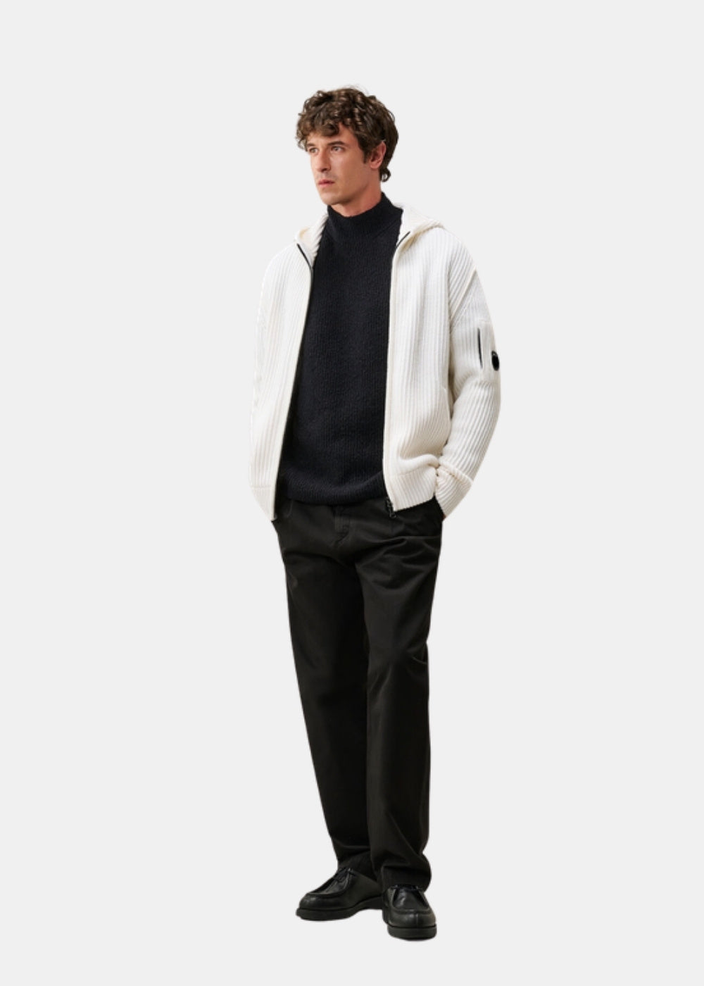 Porté en pied d'un pull à capuche zippé blanc C.P. Company associé à un pantalon sombre et des chaussures noires.