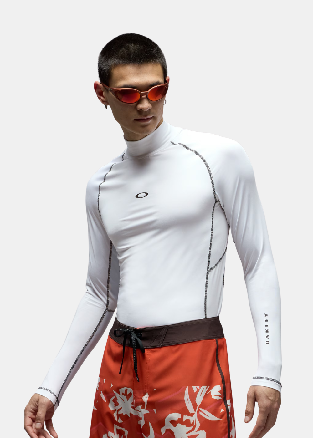 Homme portant un rashguard blanc Oakley à manches longues avec un short de bain rouge à motifs fleurs.