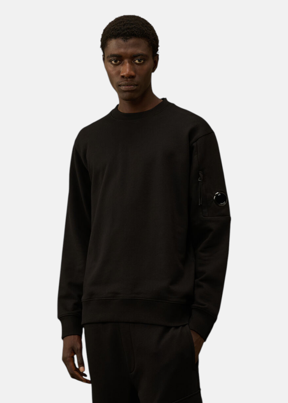 Homme portant le sweat C.P. Company col rond noir, badge lentille sur la manche, coupe regular.