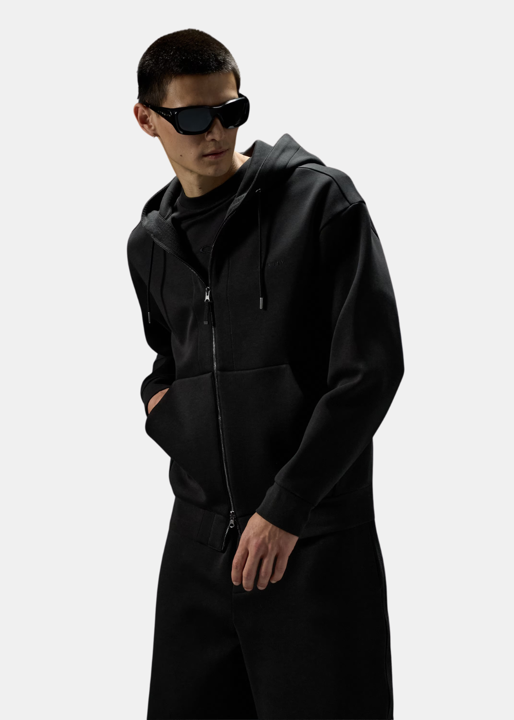 Homme portant le sweat zippé noir Oakley avec lunettes de soleil, vue de trois-quarts mettant en avant la coupe.