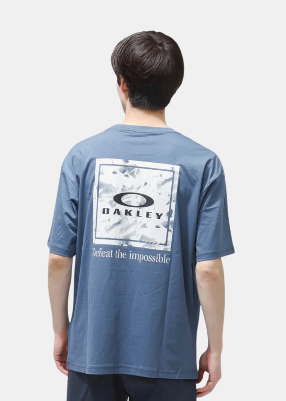 Porté de dos d'un t-shirt bleu Oakley à coupe droite montrant le grand imprimé cadre Defeat the Impossible.
