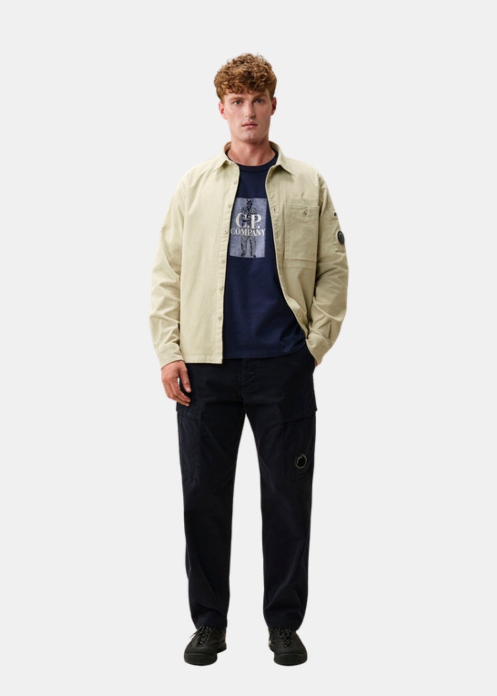 Porté en pied d'un t-shirt marine C.P. Company sous une surchemise beige et un pantalon sombre, silhouette complète.