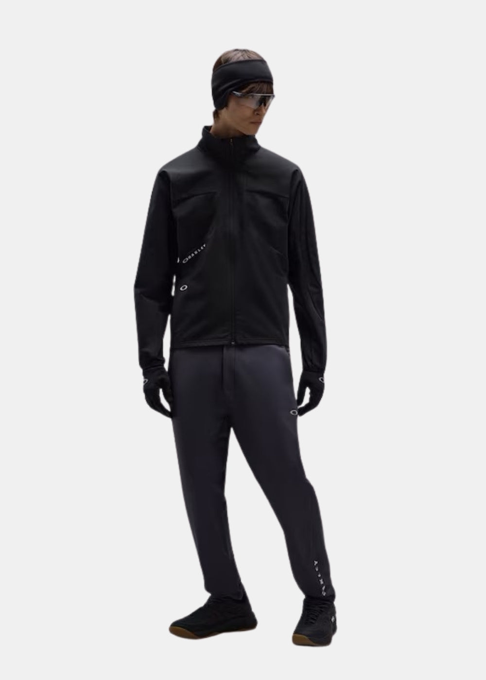 Porté en pied d'une veste technique noire Oakley Enhance avec pantalon gris anthracite, bonnet noir, gants et chaussures sombres.